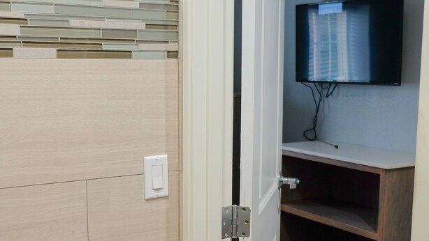 A room with a wooden floor, a white door, a mounted TV, and a shelf with a flat-screen TV.