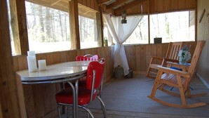Une cabane en bois avec une table ronde, des chaises rouges, un fauteuil à bascule et une fenêtre avec un rideau.
