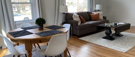 A modern living room with a wooden dining table, white chairs, a grey rug, a brown sofa with cushions, and a coffee table.