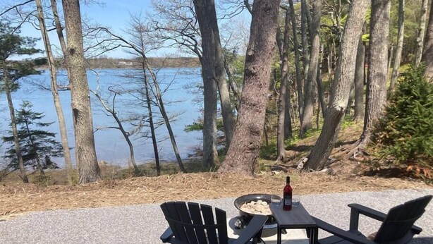 An outdoor seating area with Adirondack chairs and a table, overlooking a lake surrounded by trees. - Luxury Waterfront Tiny House (Harpswell)