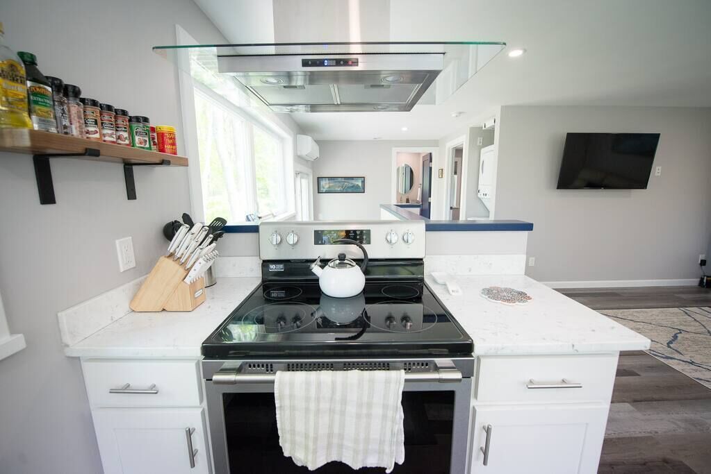 Eine moderne Küche mit Herd, Backofen und einer Dunstabzugshaube aus Glas. Es gibt Schränke, einen Messerblock und einen an der Wand befestigten Fernseher.