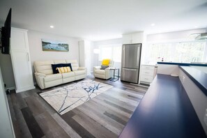 A modern kitchen with a refrigerator, a dining area with a table and chairs, and a living room with a sofa and a rug. - Stylish Treetop Loft with Balcony and Ocean Views (Harpswell)
