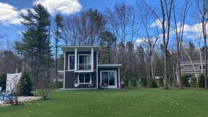 A modern two-story house with a large front lawn and surrounding trees. - Modern Cottage with Ocean Access & Wildlife (Harpswell)