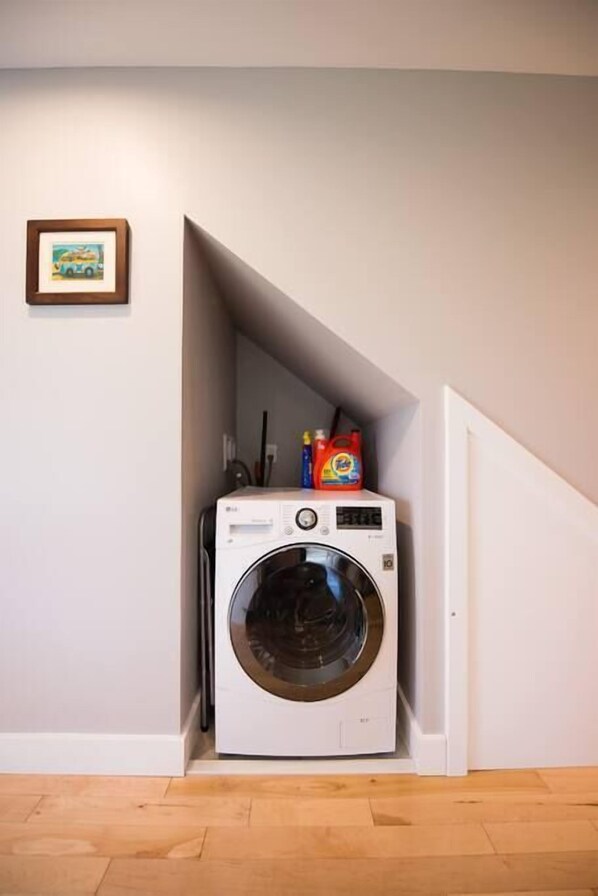 Un petit espace buanderie comprenant un lave-linge, un tableau encadré accroché au mur et des produits de lessive rangés sur le dessus.
