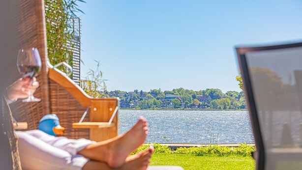 Eine Person entspannt in einem Sessel am Fenster mit Blick auf einen See, ein Laptop steht auf einem Tisch in der Nähe.