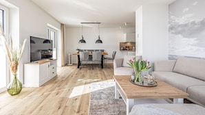 Uma sala de estar moderna com um sofá, mesa de centro e televisão. Há uma área de jantar com mesa e cadeiras. O ambiente possui piso de madeira e uma janela grande.