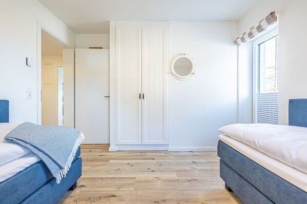 Een slaapkamer met twee bedden, een garderobe, een ronde spiegel en een raam met jaloezieën.