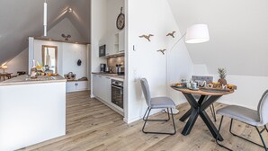 Uma cozinha moderna com armários brancos, uma área de jantar com mesa de madeira e duas cadeiras cinzas, e um quarto no mezanino com teto inclinado.