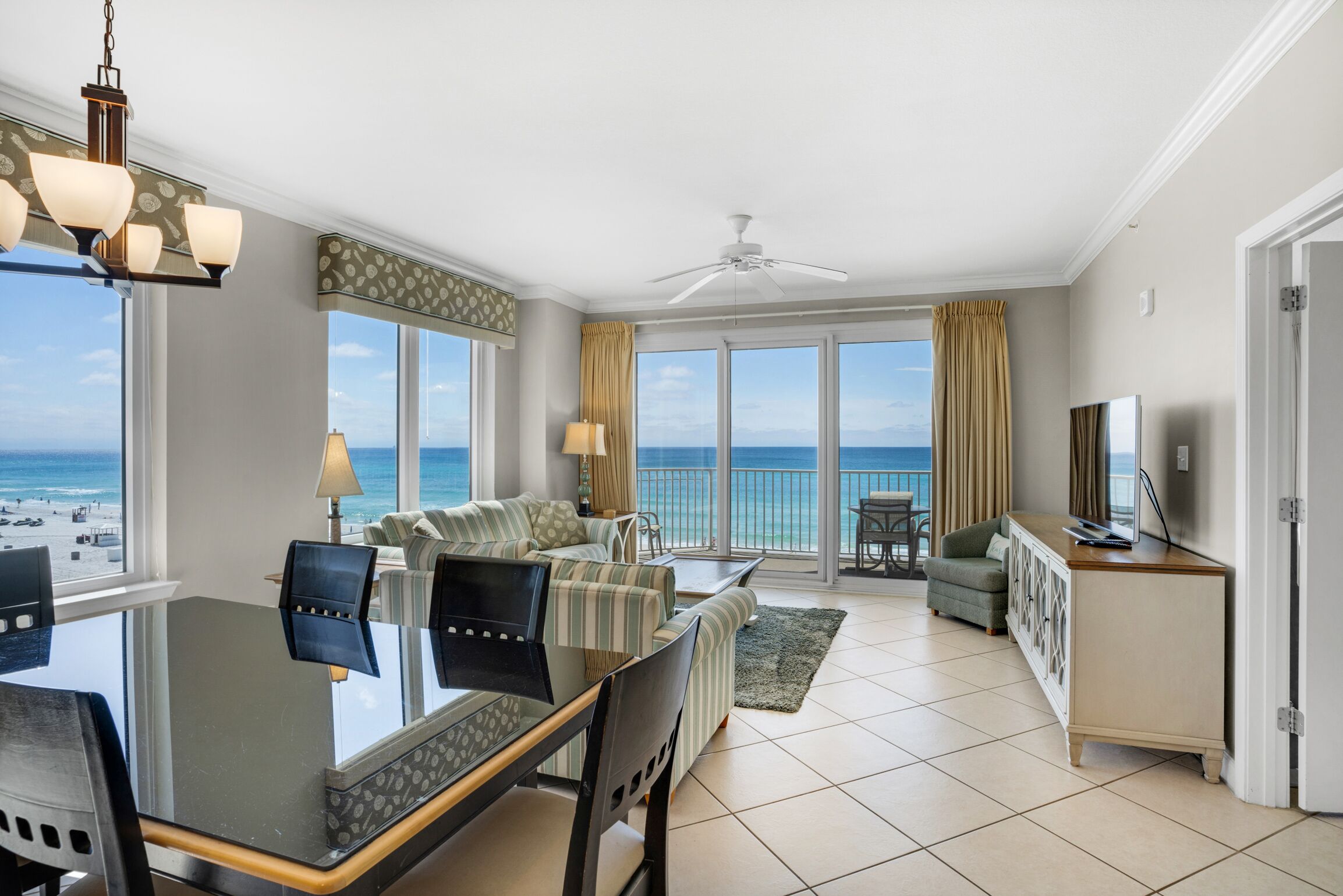 A spacious living room with a large window overlooking the ocean, a dining area with a glass table, and a balcony with a view of the beach.