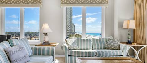 A modern living room with striped sofas, a wooden coffee table, and large windows offering a view of the beach and cityscape.