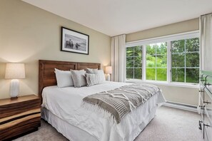 Quarto com uma grande janela, cama com colcha branca, cabeceira de madeira e mesa de cabeceira com abajur.