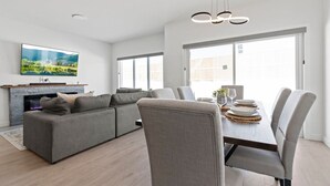 Una sala de estar moderna con un sofá seccional gris, una mesa de comedor con sillas, un televisor de pantalla plana montado en la pared y una chimenea.