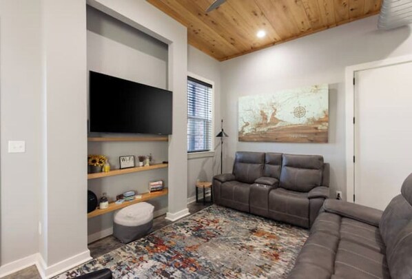 A living room with a gray sofa, a TV mounted on the wall, a wooden shelf with various items, and a colorful rug. - Historic Pensacola, a new warehouse inspired home. (Pensacola)