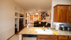 Una cocina con encimeras de granito, armarios de madera y lavavajillas. Hay una zona de comedor con un sofá y un ventilador de techo.