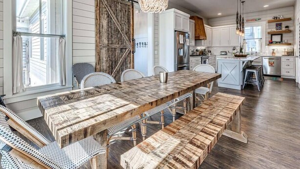 Un coin repas rustique avec une table et des bancs en bois, un lustre et, en arrière-plan, une vue sur la cuisine.