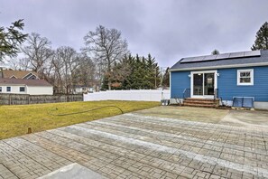 Een achtertuin met een betegeld terras, een blauw huis, een witte schutting en een houten terras.