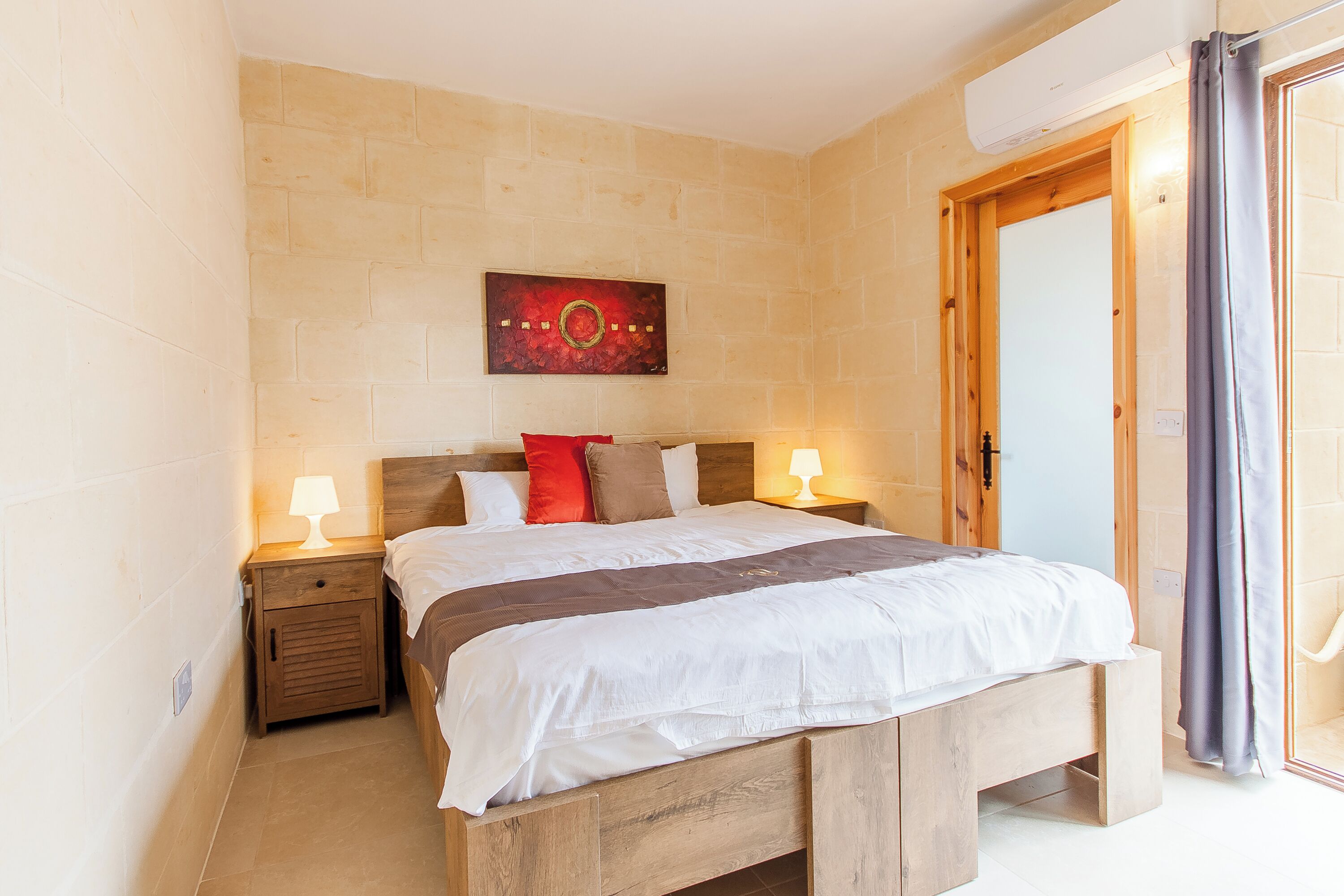 Une chambre à coucher avec un lit en bois, deux tables de chevet avec des lampes, un oreiller rouge et une œuvre d’art accrochée au mur.