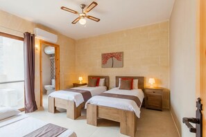 Habitación de hotel con dos camas, ventilador de techo, baño con inodoro y ventana con cortinas.