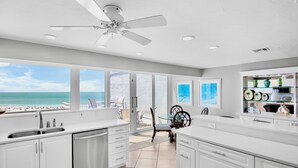 Une cuisine moderne avec des armoires blanches, un lave-vaisselle et un ventilateur de plafond. De grandes fenêtres offrent une vue sur la plage et l’océan.