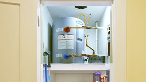 Une petite salle de bain équipée d’un chauffe-eau, de toilettes, d’un seau bleu et de produits de nettoyage.