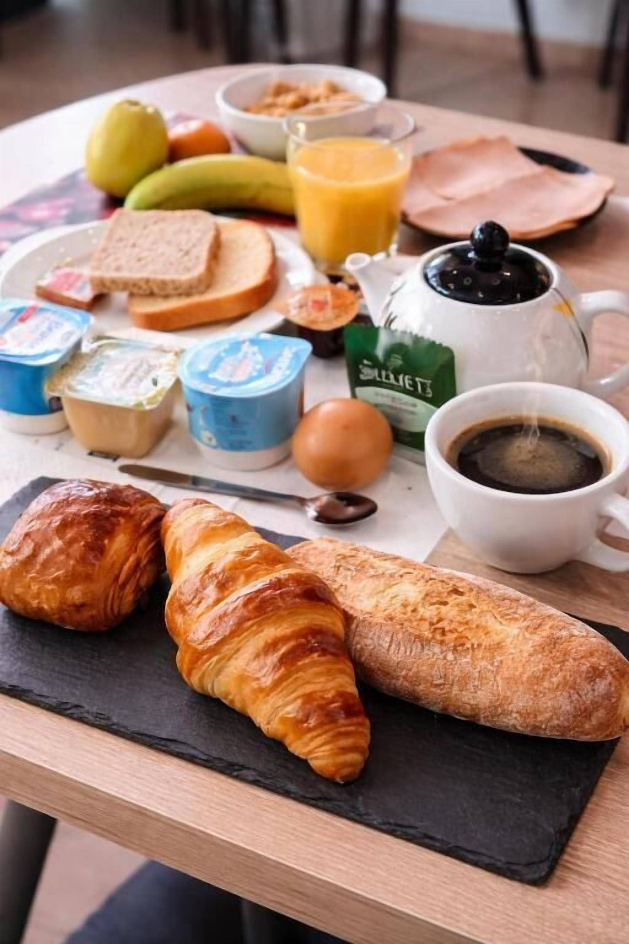 Una mesa de desayuno con cruasanes, pan, fruta, yogur y café.