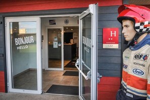 Entrada de um hotel com porta de vidro, placa de boas-vindas e um manequim usando capacete de corrida.