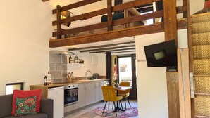 Een ruime woonkamer met een bank, een eettafel met stoelen, een keuken met moderne apparatuur, en een slaapkamer op entresol met een houten plafond.