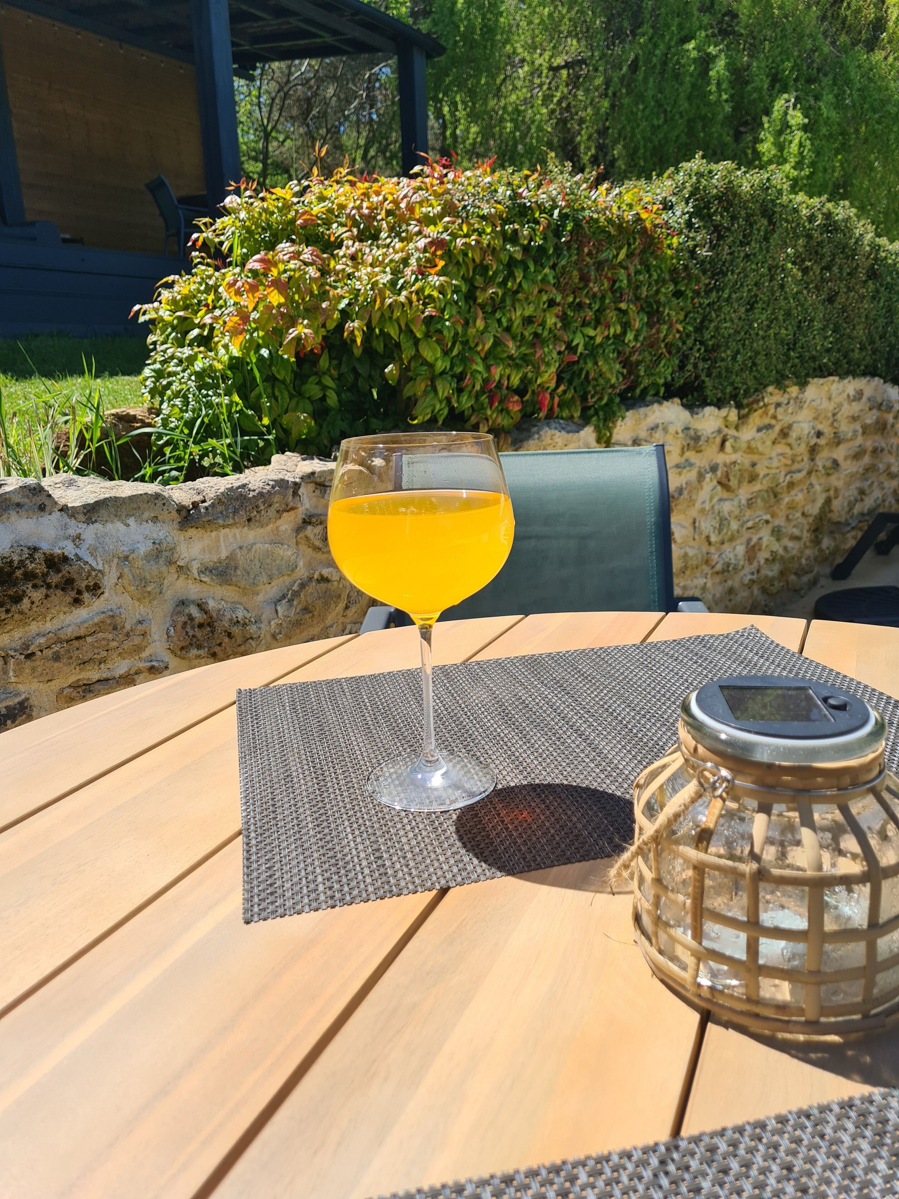 A glass of orange juice on a table with a woven placemat, a lantern, and a stone wall in the background.