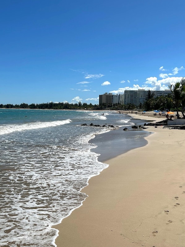 Ein Strand mit klarem, blauem Wasser, einem sandigen Ufer und einem Gebäude im Hintergrund.