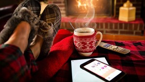 Un ambiente accogliente all'interno con un caminetto, una tazza di bevanda calda, ciabatte, un telecomando e uno smartphone su una coperta rossa.