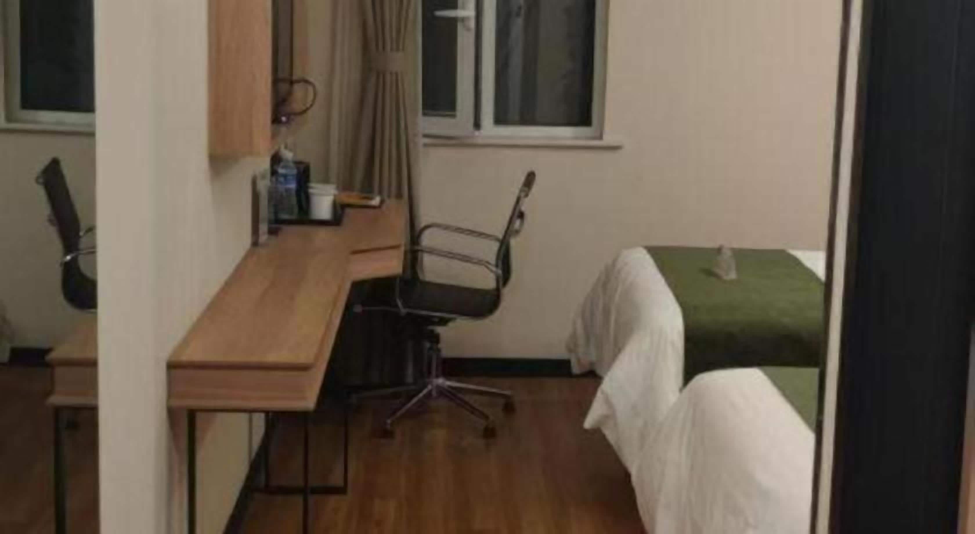 Une pièce avec un bureau en bois, une chaise, un lit recouvert d’une couverture verte et une fenêtre avec des rideaux.