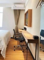 Quarto de hotel com cama, escrivaninha, cadeira, ar condicionado e janela com cortinas.