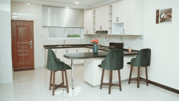 Une cuisine moderne avec des meubles blancs, un îlot central doté de tabourets de bar et une porte en bois.