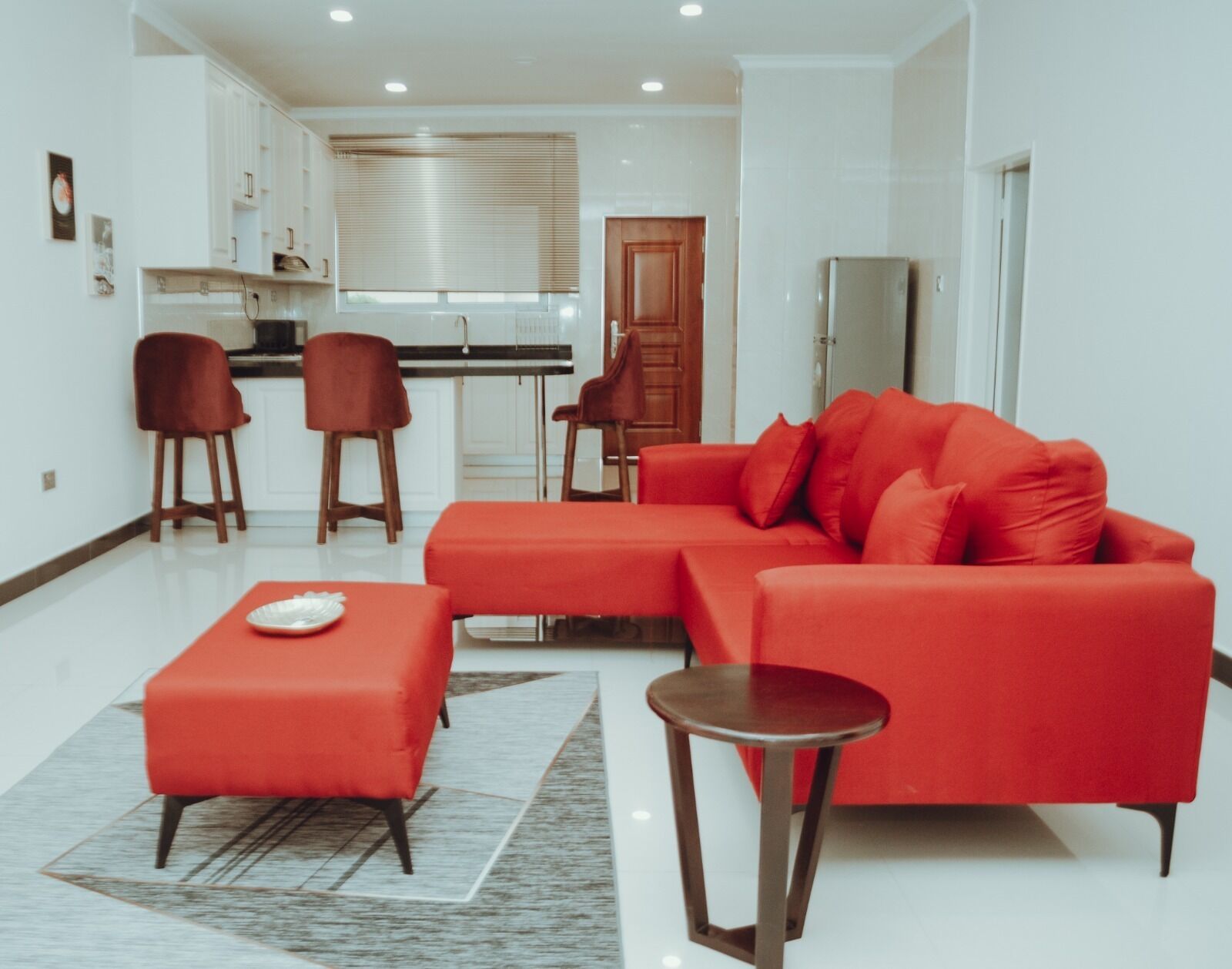 A modern living room with a red sofa, two matching armchairs, and a small table. A kitchen with white cabinets and a wooden door is visible in the background.