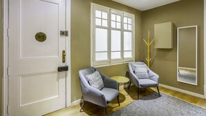 A room with a white door, two grey armchairs, a small round table, a window with white frames, a mirror, and a wooden coat rack. - Aribau D3 (Barcelona)