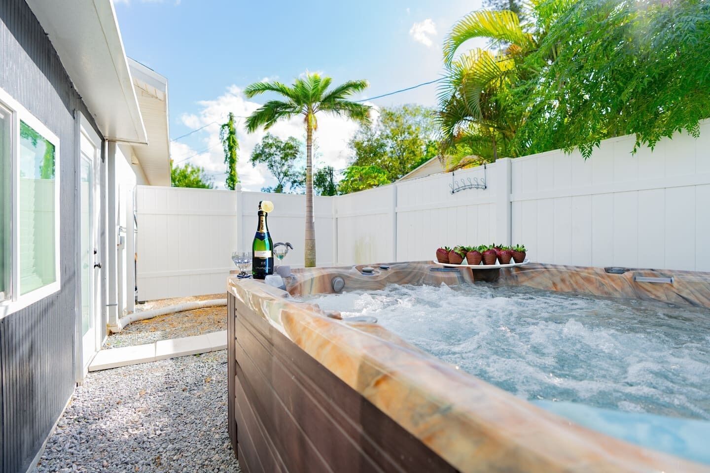 Un bain à remous avec une bouteille de champagne, entouré d’une clôture blanche, de palmiers et d’une maison aux grandes fenêtres.
