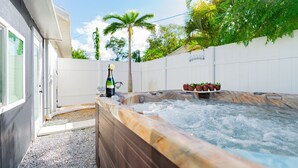Un bain à remous avec une bouteille de champagne, entouré d’une clôture blanche, de palmiers et d’une maison aux grandes fenêtres.