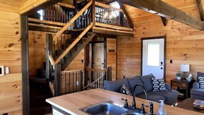 A wooden cabin interior with a kitchen area featuring a double sink, a staircase leading to a loft, and a living area with a sofa and a lamp.