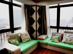 Un canapé d’angle avec des coussins verts et des motifs floraux, un panneau mural géométrique noir et blanc, et une vue sur la ville à travers de grandes fenêtres.