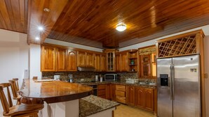 A kitchen with wooden cabinets, a stainless steel refrigerator, granite countertops, and a wooden dining table with chairs. - Villa Del Sol - Deluxe Villa 3 (Nosara)