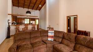 Ampla sala de estar com um sofá seccional marrom grande, teto de madeira e uma cozinha ao fundo.