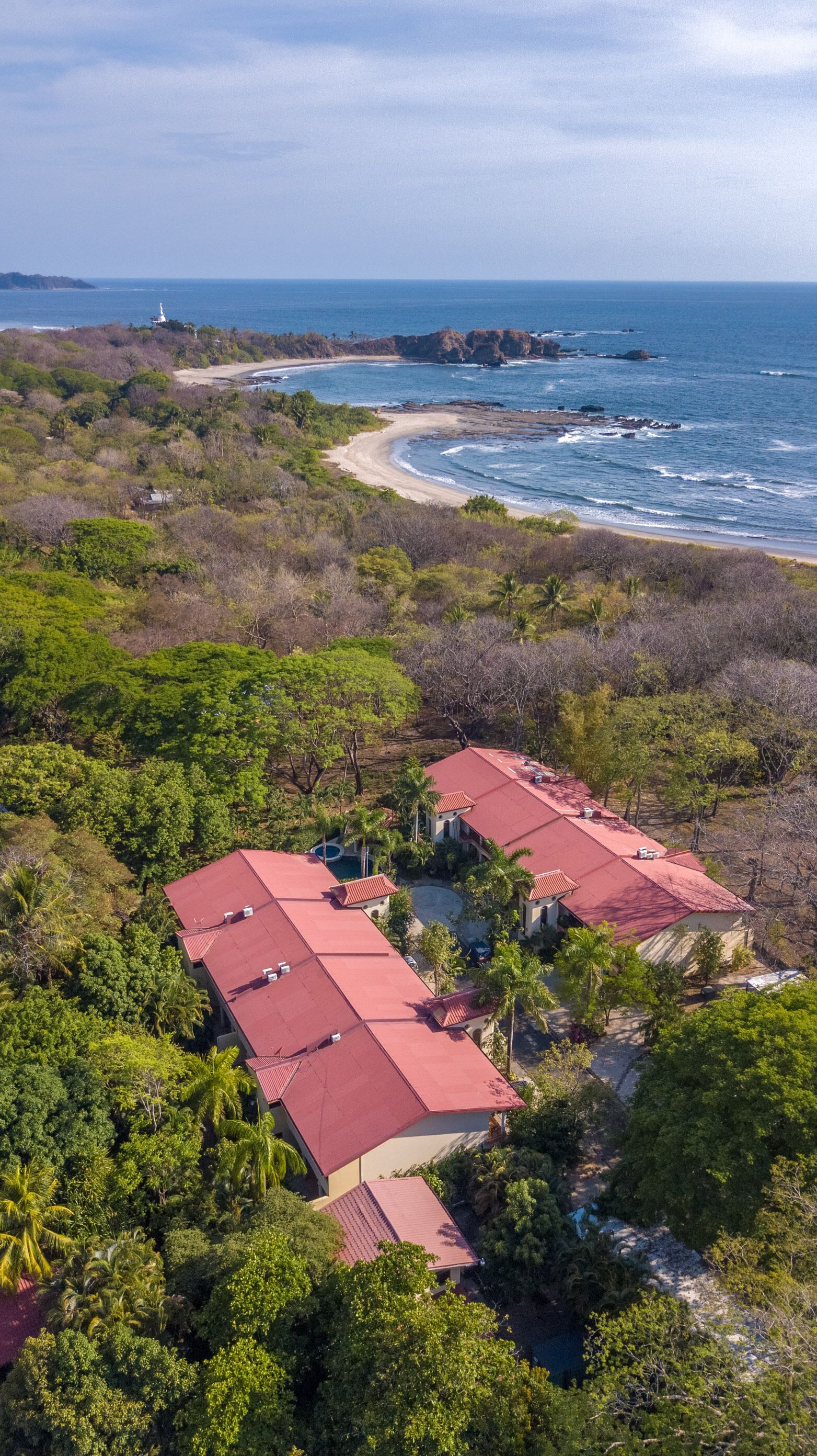 Vista aérea de un complejo turístico con varias edificaciones de techos rojos, rodeado de abundante vegetación y una playa con olas.