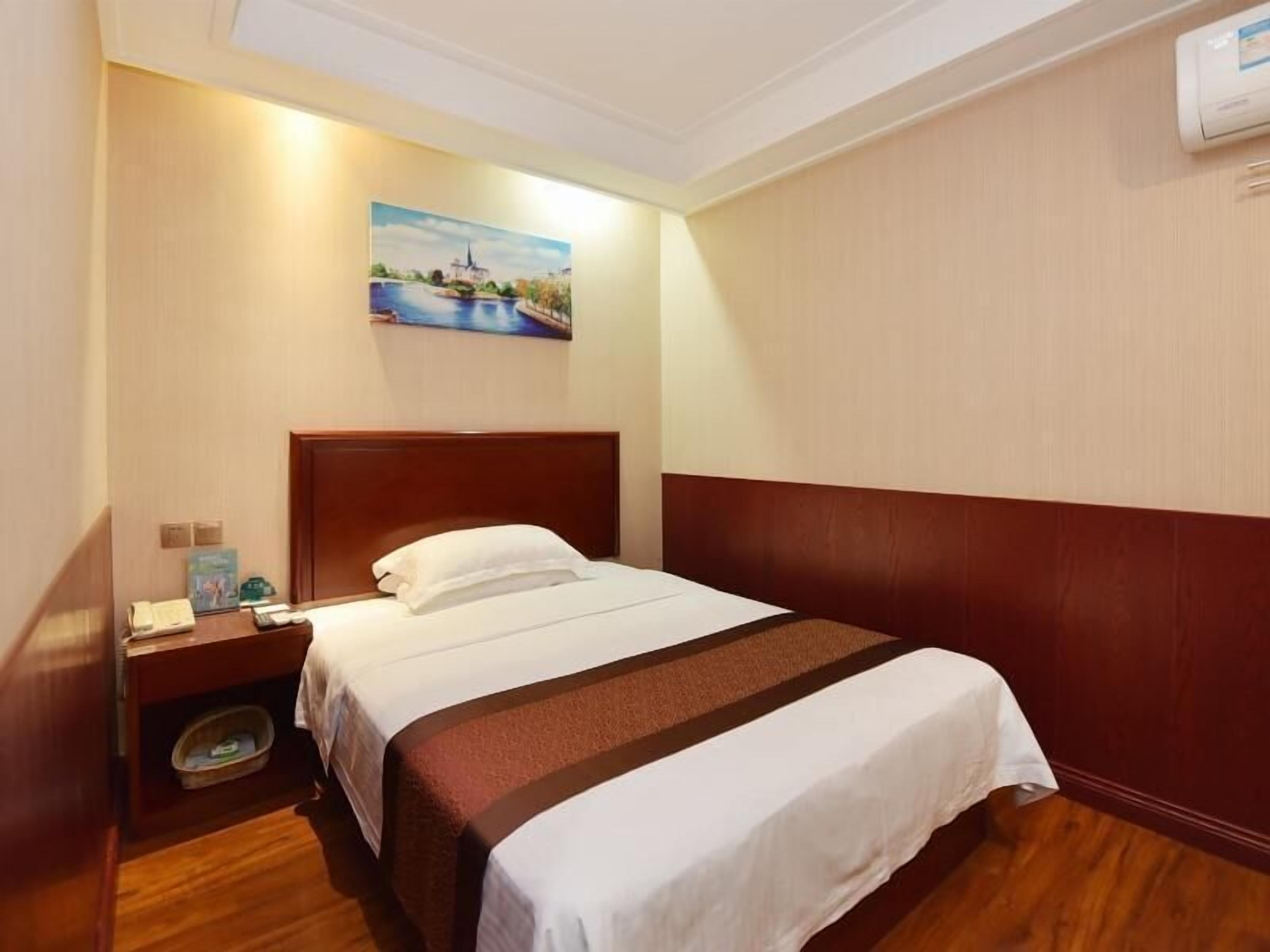 A hotel room with a wooden headboard, a bed with white bedding, a nightstand with a phone and a book, a painting on the wall, and an air conditioning unit.