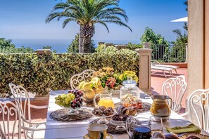 Una mesa lista para el desayuno con diversas frutas, pasteles y bebidas, con vista a un paisaje que incluye una palmera y el mar.