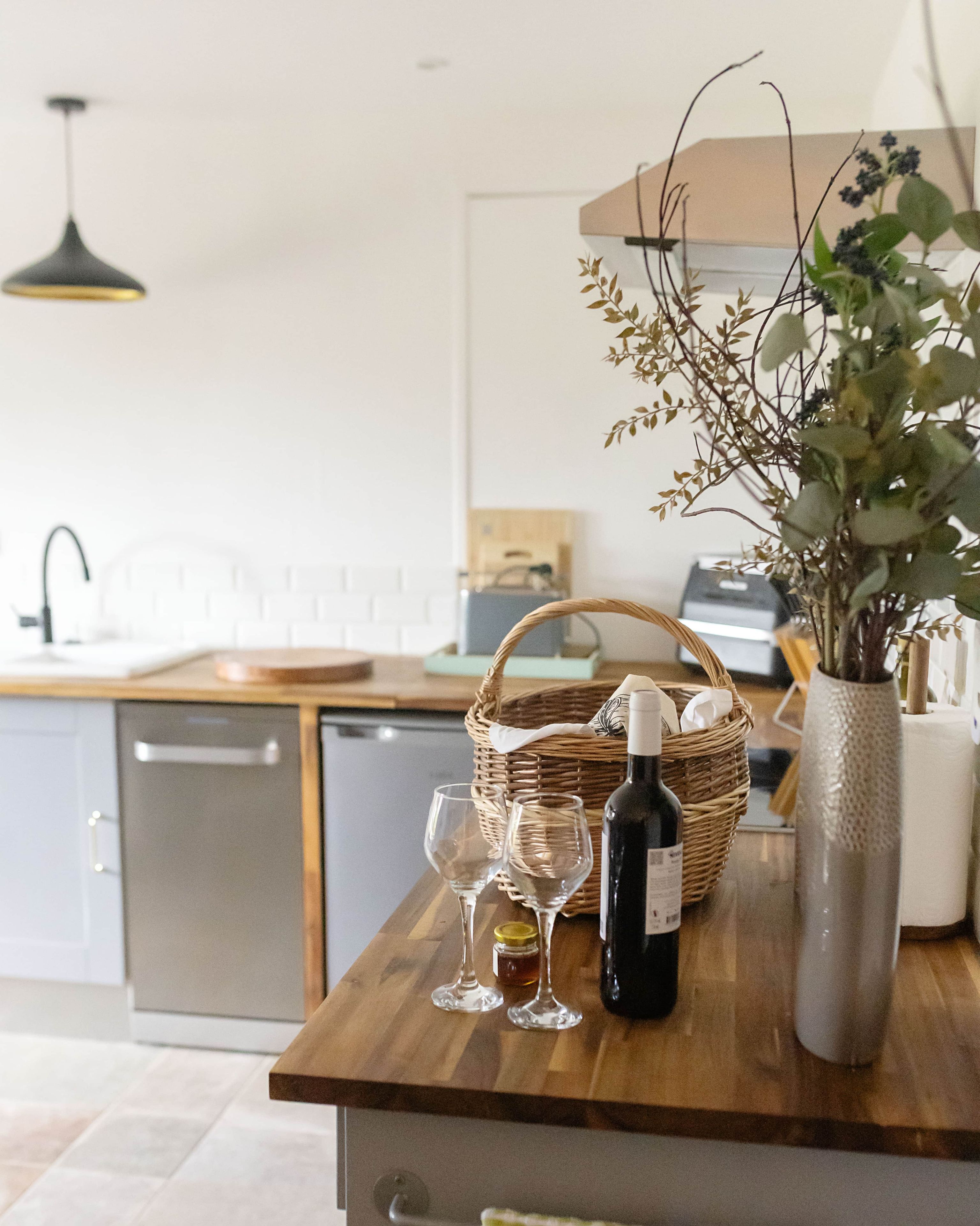 Uma bancada de cozinha com uma garrafa de vinho, duas taças, uma cesta e um vaso com folhagem.