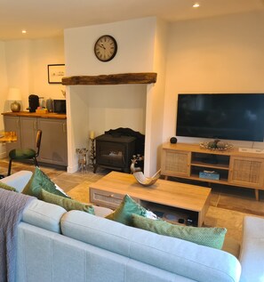 A living room with a sofa, coffee table, fireplace, television, and kitchen area. - La Prairie Gîte (Cassagnes)