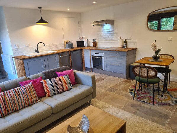 A modern kitchen with grey cabinets, a wooden countertop, and a built-in oven. There is a dining area with a wooden table and chairs, and a sofa with colorful cushions. - La Prairie Gîte (Cassagnes)