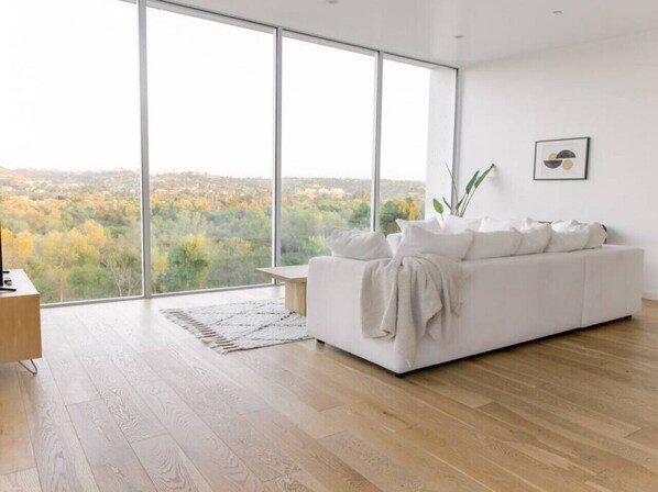 A modern living room with a large window, a white sofa, a wooden coffee table, and a plant.