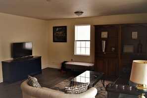A living room with a sofa, a coffee table, a TV on a cabinet, a window, and a bookshelf. - The Gateway: experience a stylish and welcoming lodging, perfect for groups. (Grand Forks)