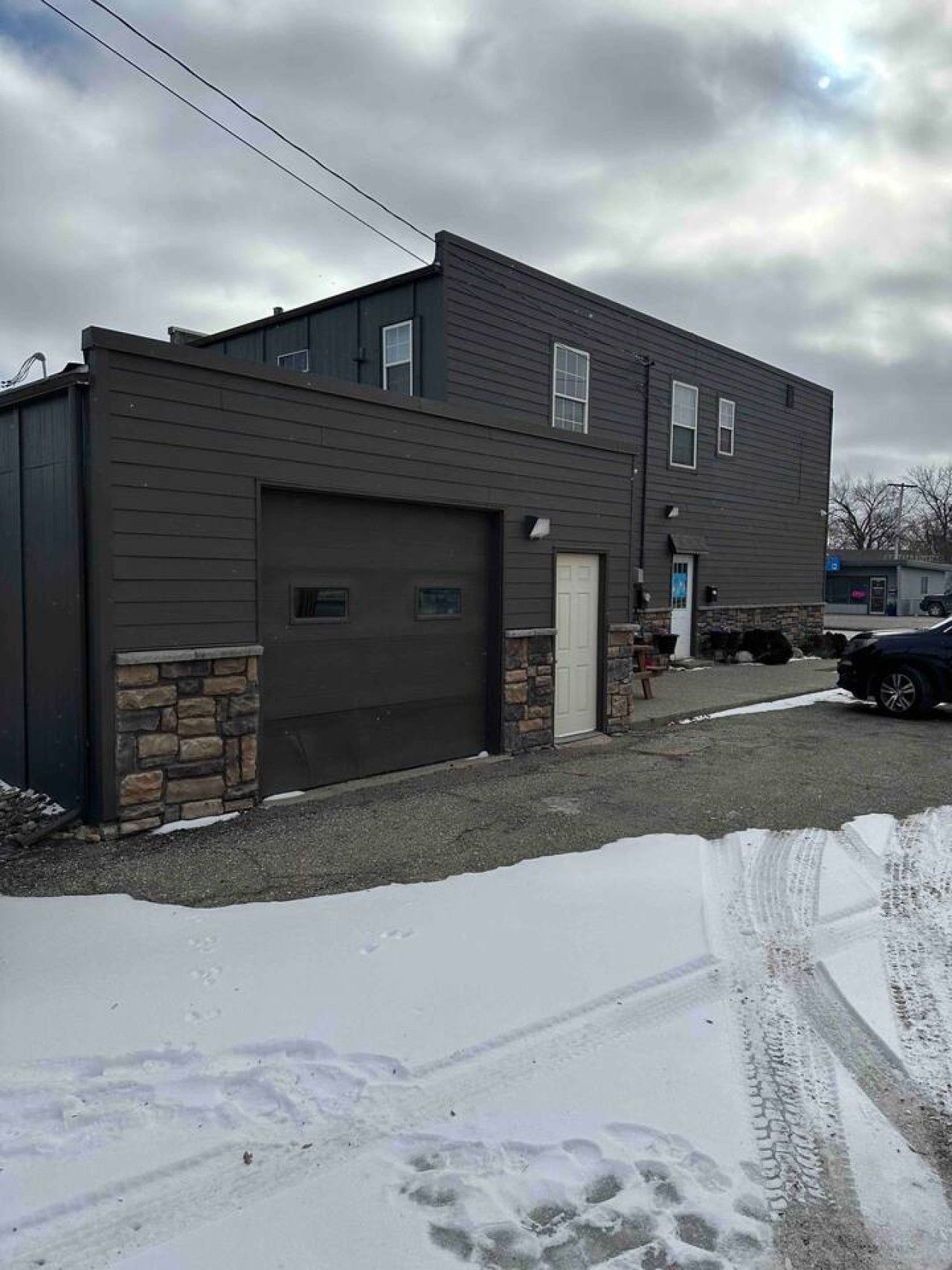 A two-story building with a garage, stone foundation, and a driveway with snow.